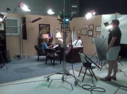 A room with a variety of lighting equipment. There are three people in the room two men and a woman sitting at a desk and working on a project. The room has a large window on the left side and a bookshelf on the right side. The floor is covered with a gray carpet and there are several lights on the walls. In the center of the image there is a large easel with a white board on it. The man standing next to the easel is wearing a black t-shirt and shorts and appears to be directing the scene. The other two people are sitting at the desk and appear to be working on the project.