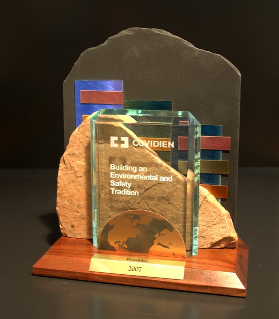 A photograph of a glass award on a wooden base. The award is rectangular in shape and has a black background. On the front of the award there is a text that reads "COVIDIEN" in white letters. Below the text it reads "Building an Environmental and Safety Tradition". On the right side of the base there are three rectangular blocks of different colors - blue red and orange. The blocks are arranged in a diagonal line with the blue block on the left and the orange block in the middle. The brown block is placed on top of a large rock which is placed next to the award. The background is black making the colors of the blocks stand out.