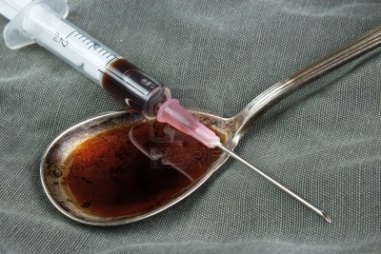 A syringe resting on a spoon that has a dark brown liquid in it likely heroin or another drug. They are resting on a gray fabric background. The needle is pointing towards the syringe as if it is about to inject it into the spoon.