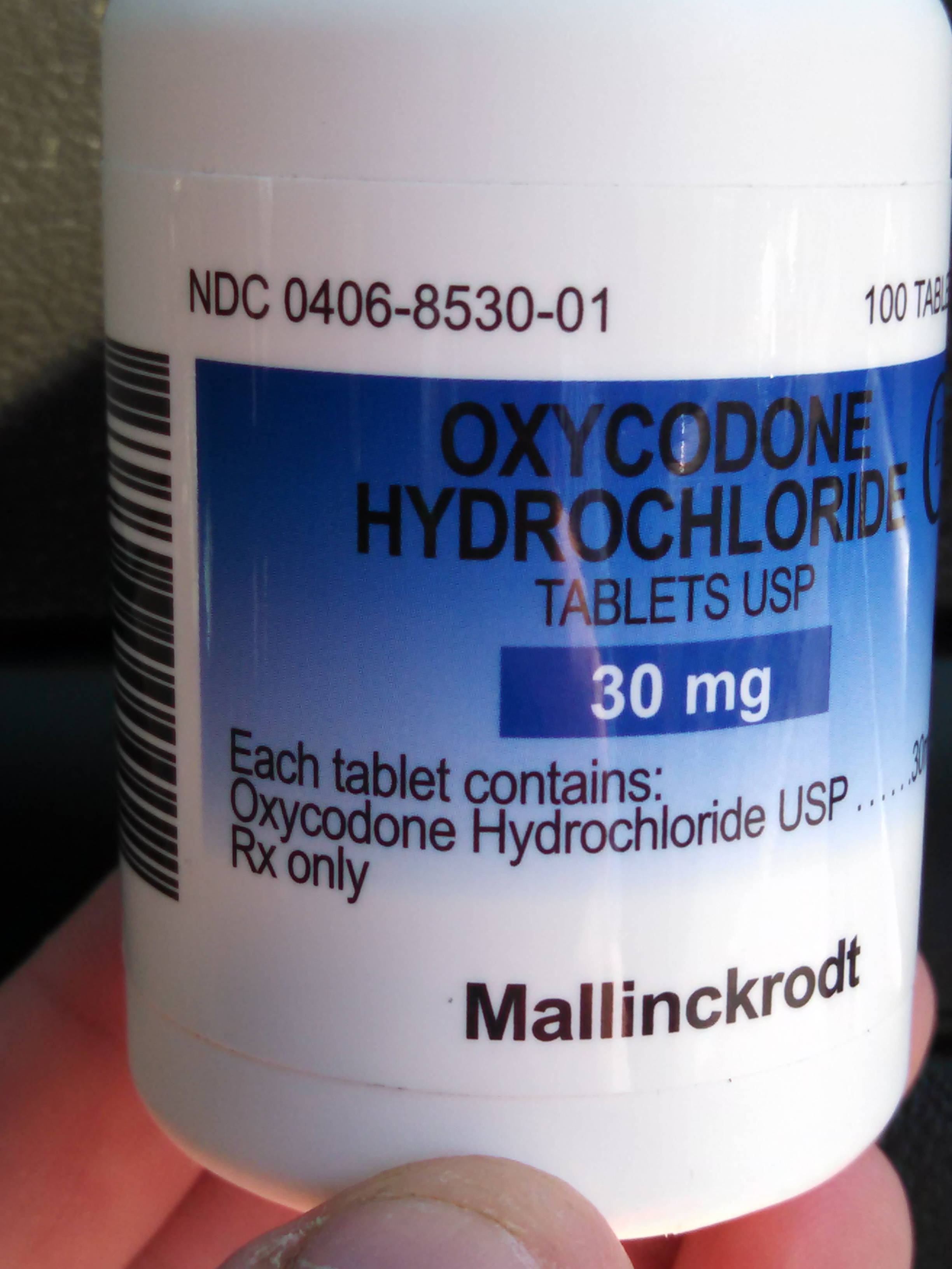 A close-up of a white pill bottle with a blue label. The label has the text "Oxycodone Hydrochloride Tablets USP 30 mg" written in bold black letters. Below the text there is a barcode and the number "NDC 0406-8530-01" in smaller black letters indicating that the bottle contains 100 tablets. The bottle is held in a person's hand and the background is blurred.