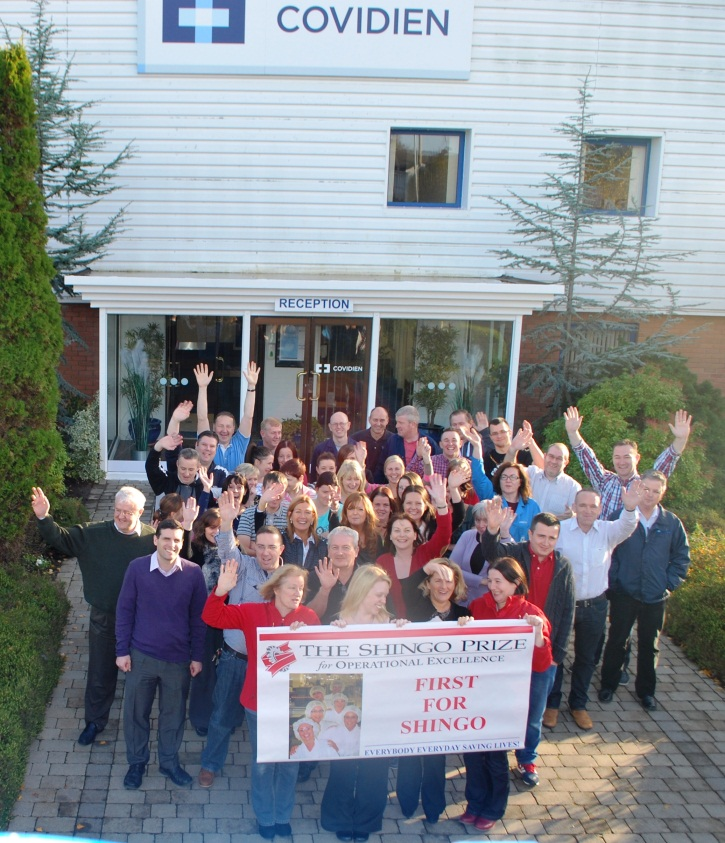 A group of people standing in front of a building with a sign that reads "Covidien". The building appears to be a hospital or clinic as there is a blue cross on the top of the building and a reception desk on the right side. The people in the group are smiling and waving their hands in the air. In the center of the group there are two women holding a banner that reads "#The Shingo Prize First for Shingo". The group is standing on a brick pathway and there are trees and bushes in the background.