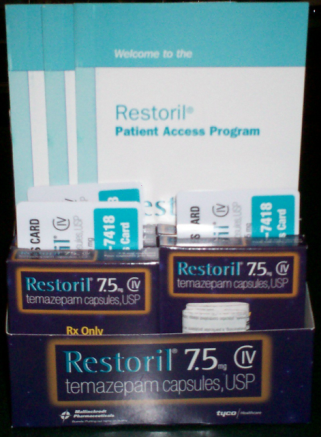 A box of Restoril 75.5 mg tablets. The box is blue and white with the brand name "Restoril" written in bold letters at the top. Below that there is a sign that reads "Welcome to the Restoril Patient Access Program". <br /><br />The box is stacked on top of each other with the top box slightly tilted to the left. The bottom box is rectangular in shape and has the brand's logo and the product name written on it. The top box has a label that says "Rx Only" and "Temazepam Capsules USP". The box also has a picture of the tablets on it which are white and blue in color.<br /><br />There are also two small white cards with the same brand name and product name on them. The cards are placed on the top left corner of the box. The background is dark making the blue box and the white cards stand out.