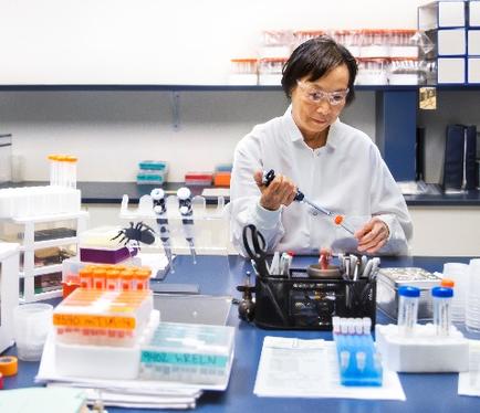 A woman in a laboratory setting. She is wearing a white lab coat and glasses and is holding a pipette in her hand. She appears to be working on a test tube experiment. On the table in front of her there are several test tubes of different sizes and colors as well as other scientific equipment. There are also several other test tubes and tubes in a black tray. In the background there is a rack with more test tubes. The woman seems to be focused on her work and is likely conducting a scientific experiment.
