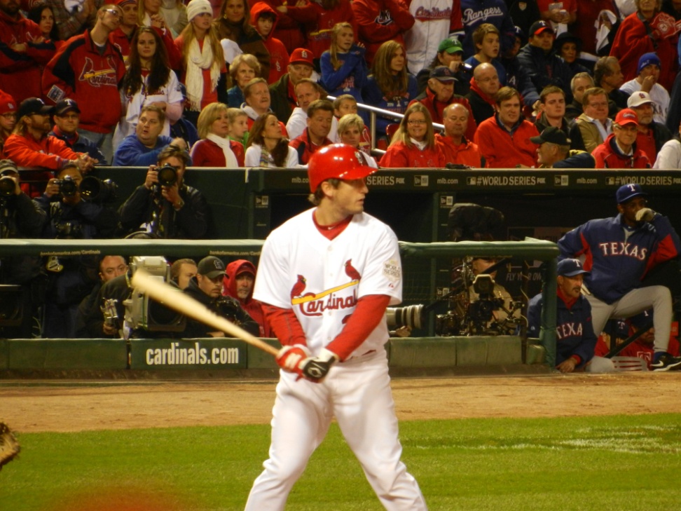 A baseball player from the St. Louis Cardinals team at bat during a game. The player is wearing a red and white uniform with the team's logo on the front and the word "Cardinals" written across the chest. He is holding a wooden bat and appears to be in the middle of a swing. The background shows a large crowd of spectators in the stands some of whom are holding cameras and taking pictures. The stands are filled with people wearing red and blue uniforms and there is a sign that reads "Texas Rangers" on the right side of the image. The field is covered in green grass and there are a few spectators visible in the background.