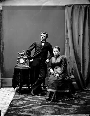 A black and white photograph of a man and a woman posing for a portrait. The man is standing on the left side of the image wearing a suit and tie with his hands in his pockets. He has a serious expression on his face and is looking directly at the camera. The woman is sitting on a chair on the right side with her legs crossed and her hands resting on her lap. She is wearing a long dress with a high neckline and long sleeves. The background is a room with a large window and a curtain and there is a small table with a clock on it in front of the man. The floor is covered with a patterned rug. The photograph appears to be from the early 20th century.