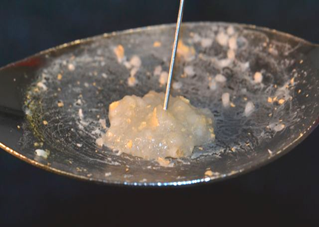 A frying pan with a small amount of food in it. The pan appears to be made of stainless steel and has a shiny surface. The food in the pan is a light yellow color and looks like it is being cooked. A metal spoon is resting on the edge of the pan and there are small pieces of garlic scattered around the pan. The background is dark making the food stand out.