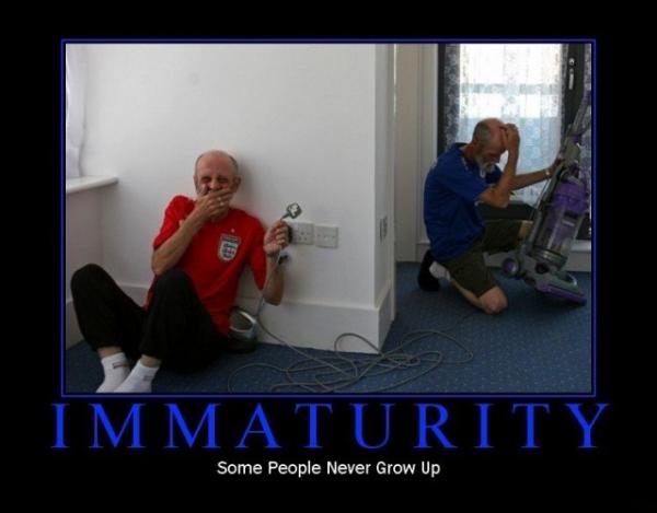 Two elderly men in a room with a vacuum cleaner. The man on the left is wearing a red t-shirt and black pants and is sitting on the floor with his hands covering his mouth. He appears to be upset or frustrated with his eyes closed and his mouth slightly open. The other man is kneeling next to him wearing a blue shirt and is holding the vacuum cleaner with both hands. The background shows a white wall and a window with blinds. The image is accompanied by text that reads "Immaturity: Some People Never Grow Up."