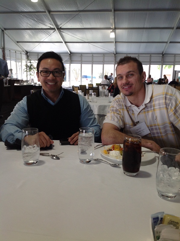 Two men sitting at a table in a restaurant. They are both smiling and appear to be enjoying their meal. The table is covered with a white tablecloth and there are two glasses of water on the table. The man on the left is wearing a black vest and glasses and is holding a phone in his hand. He is also smiling and looking at the camera. On the right side of the table there is a plate of food and a glass of soda. In the background there are other tables and chairs with people sitting at them. The restaurant has a high ceiling with white walls and a large window that lets in natural light.