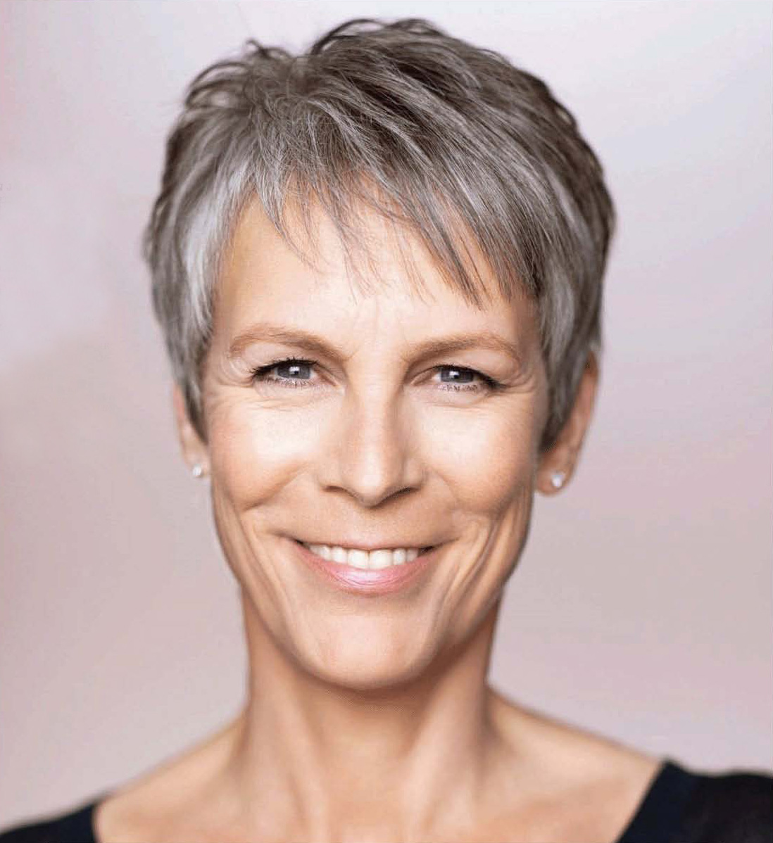 A close-up portrait of a middle-aged woman with short gray hair. She is smiling widely and looking directly at the camera. Her hair is styled in a short layered cut with bangs framing her face. She has a slight smile on her lips and her eyes are bright and full of life. She appears to be in her late 40s or early 50s. The background is a neutral grey color making the woman the focal point of the image.