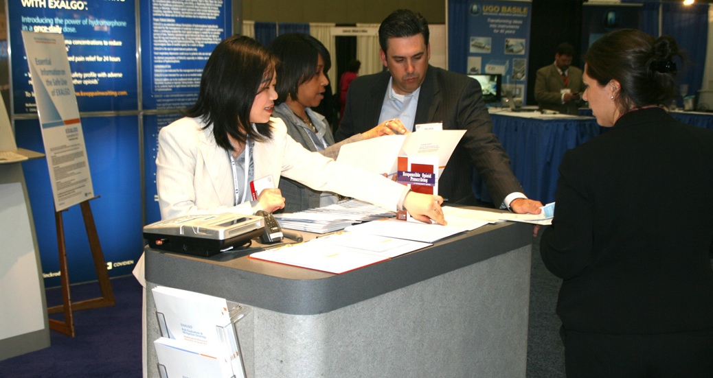 A group of people at a trade show booth. There are three people in the image two women and one man standing behind a reception desk. The woman on the left is a young woman with long dark hair wearing a white blazer and a name tag. She is holding a piece of paper in her hand and appears to be explaining something to the man on the right who is wearing a suit and tie. The man is standing behind the desk and is looking at the papers. There is a blue banner in the background with white text and images on it. The booth is set up in a large room with blue walls and a blue carpet.