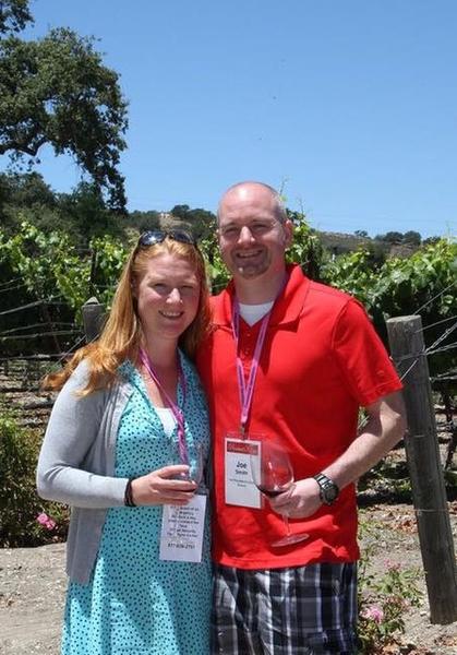 A man and a woman standing in a vineyard. The man is wearing a red polo shirt and plaid shorts and is holding a glass of red wine in his hand. He is also wearing a lanyard around his neck. The woman is standing next to him wearing a blue dress and a gray cardigan. They are both smiling and looking at the camera. In the background there are rows of grapevines and a clear blue sky.