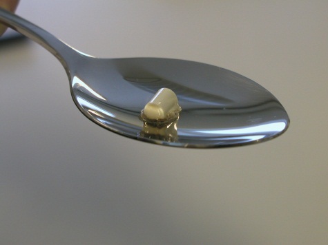 A close-up of a silver spoon with a small amount of white substance on it likely heroin or another drug. The background is a solid gray color and the focus is on the spoon.