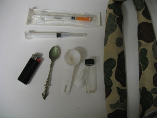 A collection of medical supplies laid out on a white surface. On the left side of the image there is a black lighter with a red cap. Next to it there are two syringes one with an orange needle and the other with a clear plastic tube. In the center of the table next to the lighter is a silver spoon with a long handle and a small white bowl with a black lid. On top of the spoon is a small glass jar with a lid. The background is a beige and brown patterned fabric.