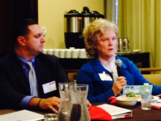 A man and a woman sitting at a table in a conference room. The man is on the left side of the image and the woman is sitting on the right side. They are both wearing name tags and appear to be engaged in a conversation. The woman is holding a microphone and speaking into it while the man is listening attentively. On the table in front of them there are plates of food a water bottle and a glass of water. In the background there is a coffee machine and a window with curtains. The room appears to be well-lit with natural light coming in from the window.