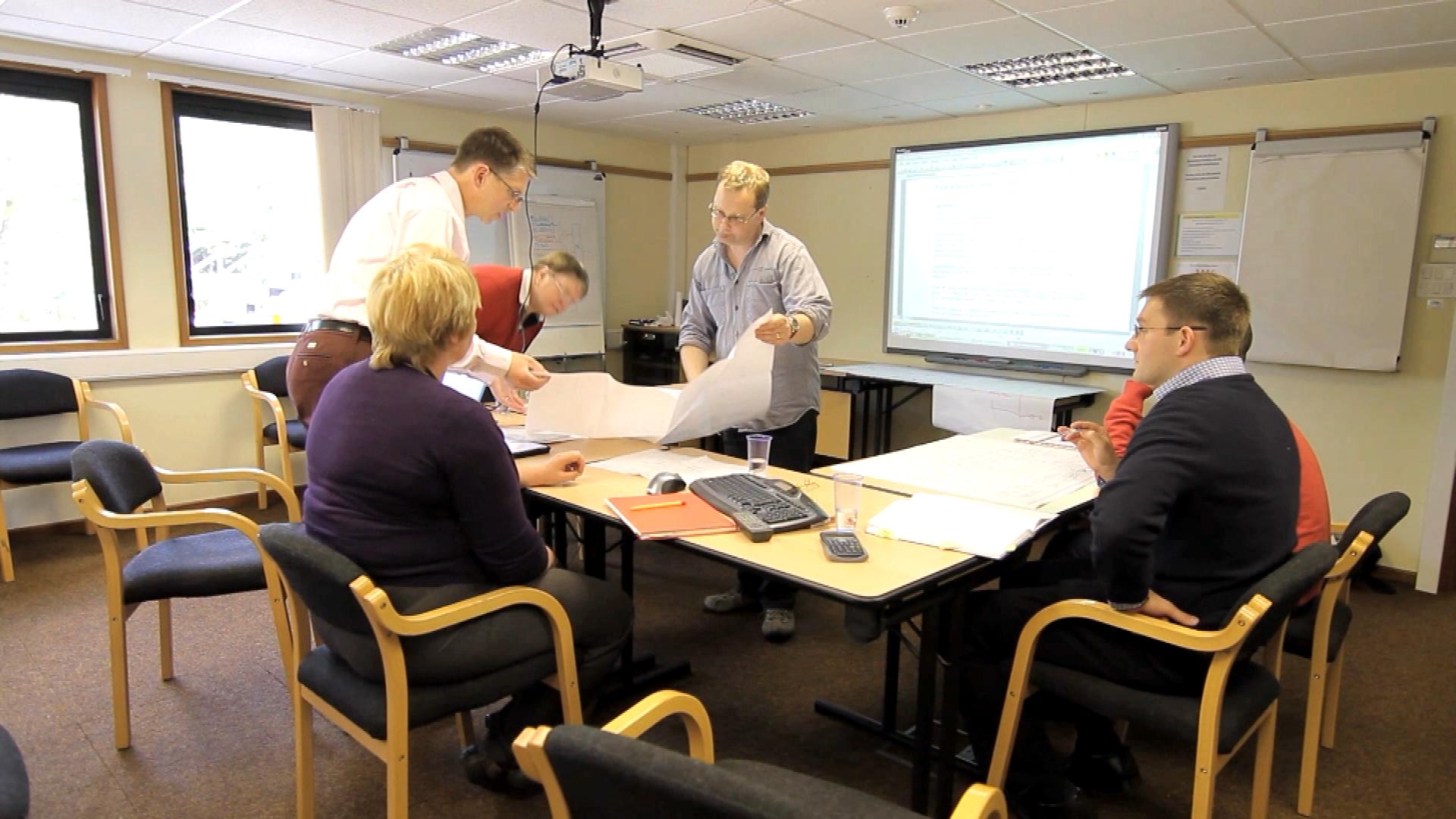 A group of five people in a meeting room. There are four people in the room three men and two women sitting around a table with papers a calculator and other office supplies scattered across it. They appear to be engaged in a discussion or brainstorming session. The room has a large window on the left side and a projector screen on the right side and a whiteboard on the wall in the background. The people are dressed in casual business attire and are looking at the papers on the table.