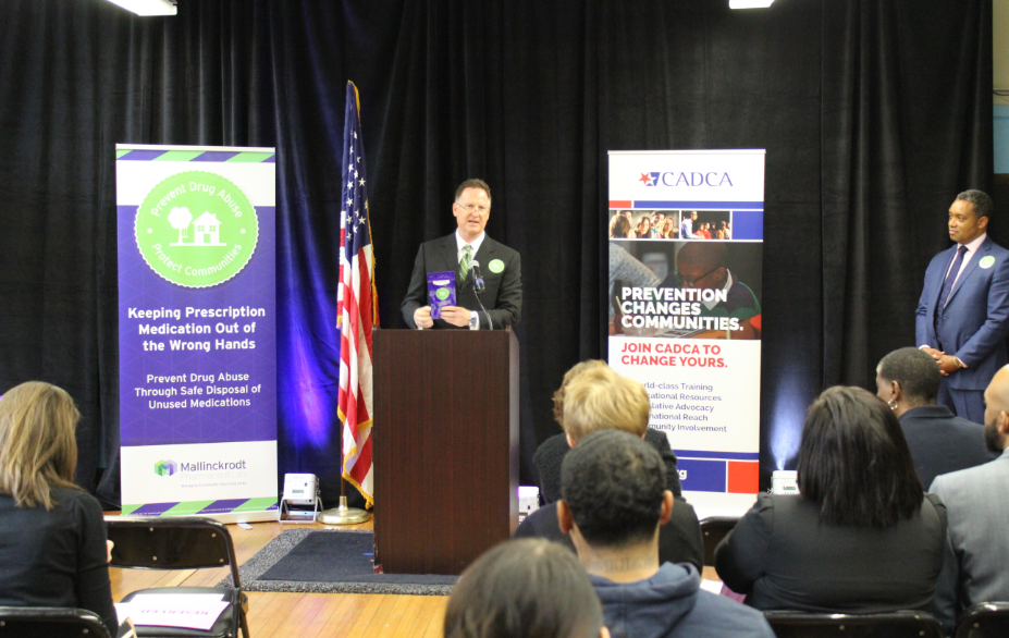 A man standing at a podium in front of a group of people sitting in chairs. He is speaking into a microphone and appears to be giving a presentation. Behind him there is an American flag and a banner that reads "Keeping Prescription Medicine Out of the Wrong Hands". On the right side of the image there are two men standing at the podium one of whom is holding a microphone. The background is a black curtain and there is a projector screen on the left side. The people in the audience are attentively listening to the presentation.