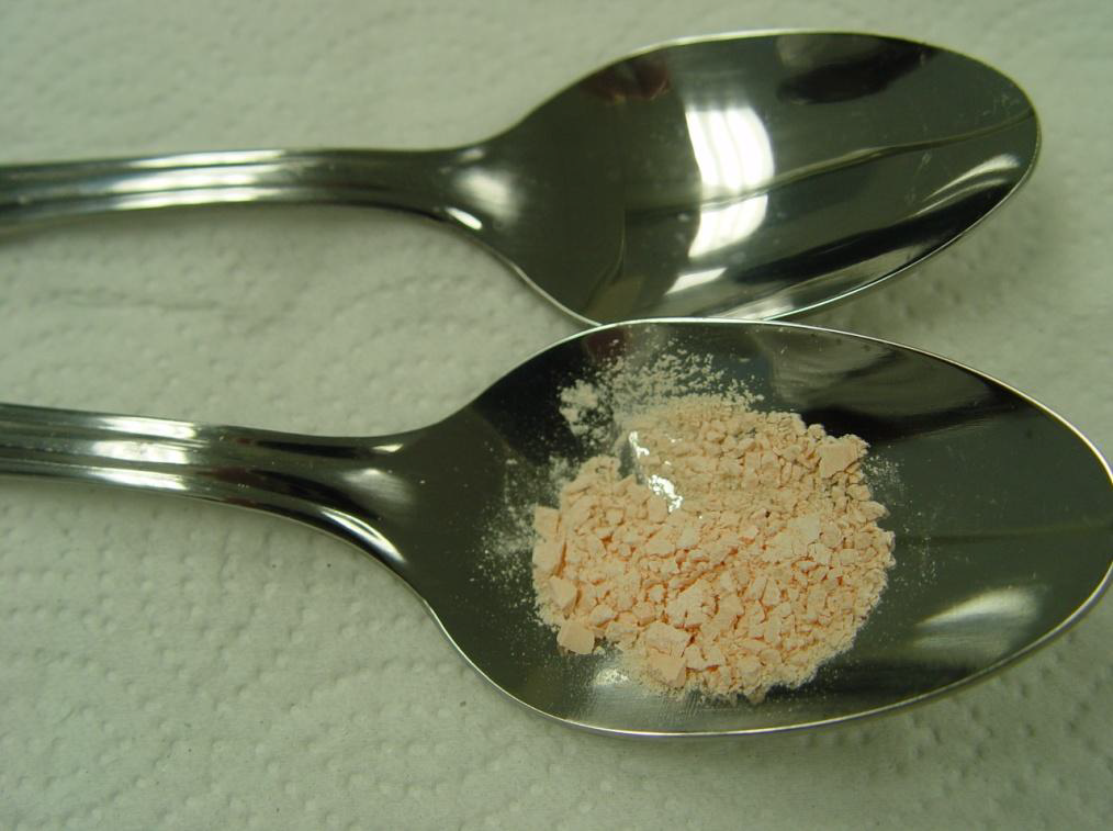 Two silver spoons lying side by side on a white surface. The spoons have a shiny finish and appear to be made of stainless steel. On one of the spoons there is a small amount of pink powder which appears to be a type of powdery substance. The powder is finely ground and has a textured surface.