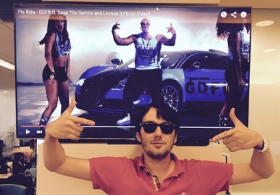 A man standing in front of a large screen with a video playing on it. The man is wearing a red polo shirt and sunglasses and has a beard. He is holding up two thumbs up in a thumbs up gesture. On the screen there is an image of a man wearing a blue tank top and black shorts standing next to a blue sports car. The background of the screen is white and there is text on the top right corner that reads "Flo Rida - The Gemini and Lookas (Official Video)".