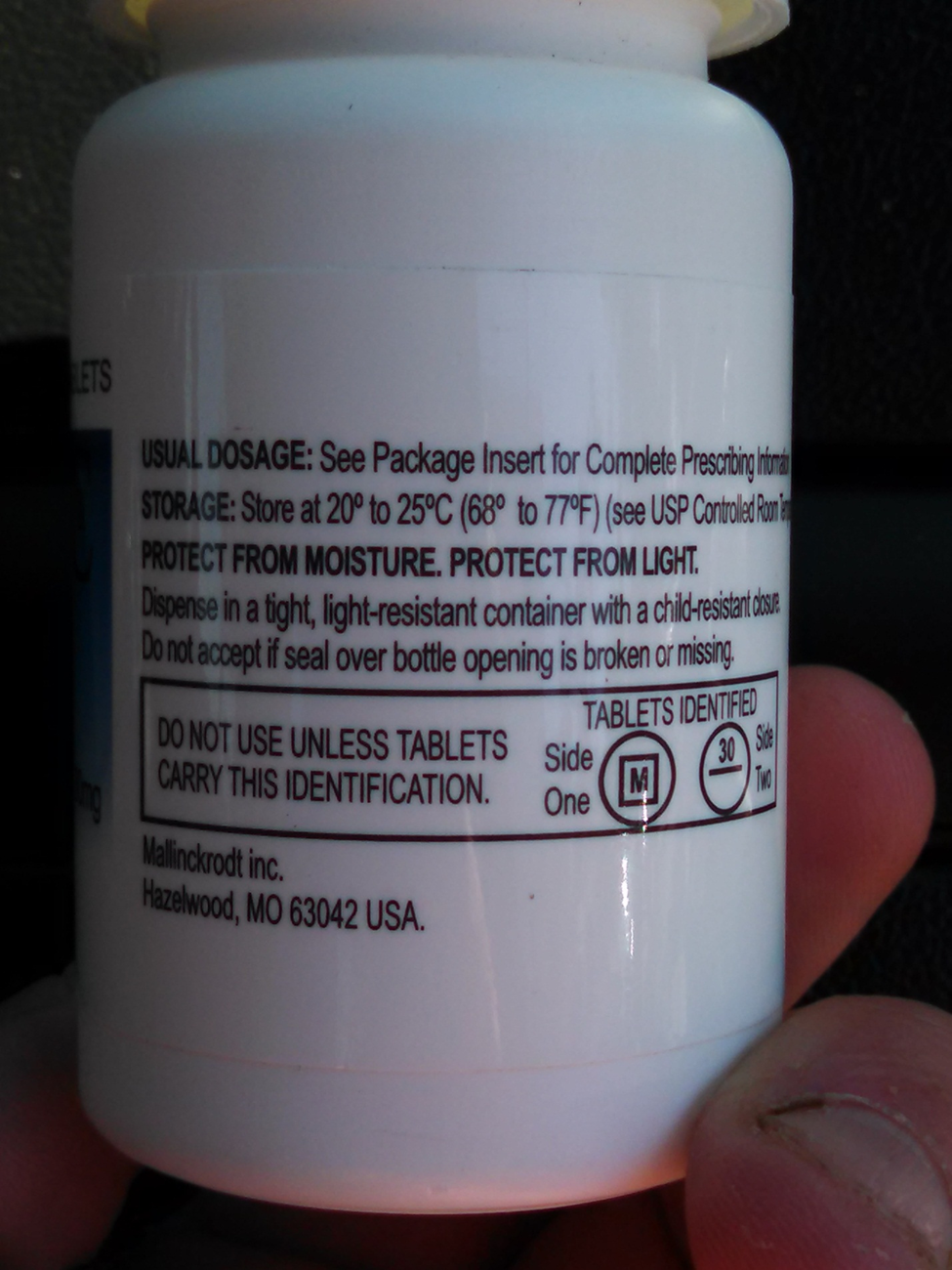 A close-up of a white pill bottle with a yellow cap. The label on the bottle is black and white and has text written on it. The text reads "Usual dosage: See Package Insert for Complete Prescription Information. Storage: Store at 20°C to 25°C (68°C) to 77°F (see USP Controlled Room). Protect from moisture. Protect from light. Dispense in a tight light-resistant container with a child-resistant closure. Do not accept if seal over bottle opening is broken or missing. Do NOT USE UNLESS TABLETS. Carry this identification. One side of the label is labeled "Tablets Identified." The label also mentions that the pill bottle contains one side of tablets. The background is dark and the bottle appears to be held in a person's hand.