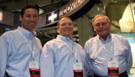 Three men standing in front of a booth with the word "COVIDIEN" written on it. They are all wearing light blue collared shirts and have name tags around their necks. The man on the left is a young man with dark hair the man in the middle is a middle-aged man with short blonde hair and the man next to him is an older man with white hair. All three men are smiling and appear to be posing for the photo. In the background there is a large screen displaying information about the booth.