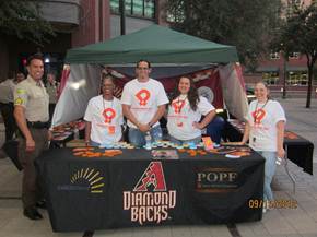 A group of five people standing behind a table with a green tent. The table is covered with a black tablecloth and has the words "Diamond Backs" and "Pope" written on it. The people are wearing white t-shirts with the same logo on them. They are smiling and appear to be posing for a photo. Behind the table there is a man in a police uniform standing next to the table. The background shows a city street with buildings and trees.