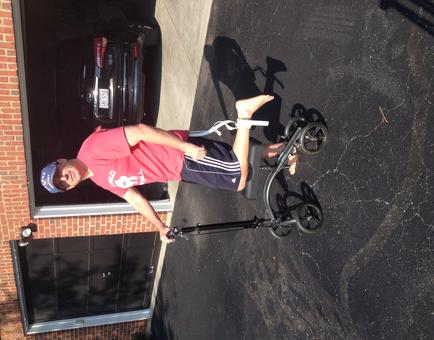 A man standing in front of a brick building with a black car parked in the background. He is wearing a red t-shirt black shorts and a blue baseball cap. He has his left leg bent at the knee and his right leg extended behind him. The man is holding onto a black tricycle with four wheels. The tricycle appears to be in good condition with no visible damage or wear. The image is taken from a low angle looking up at the man.