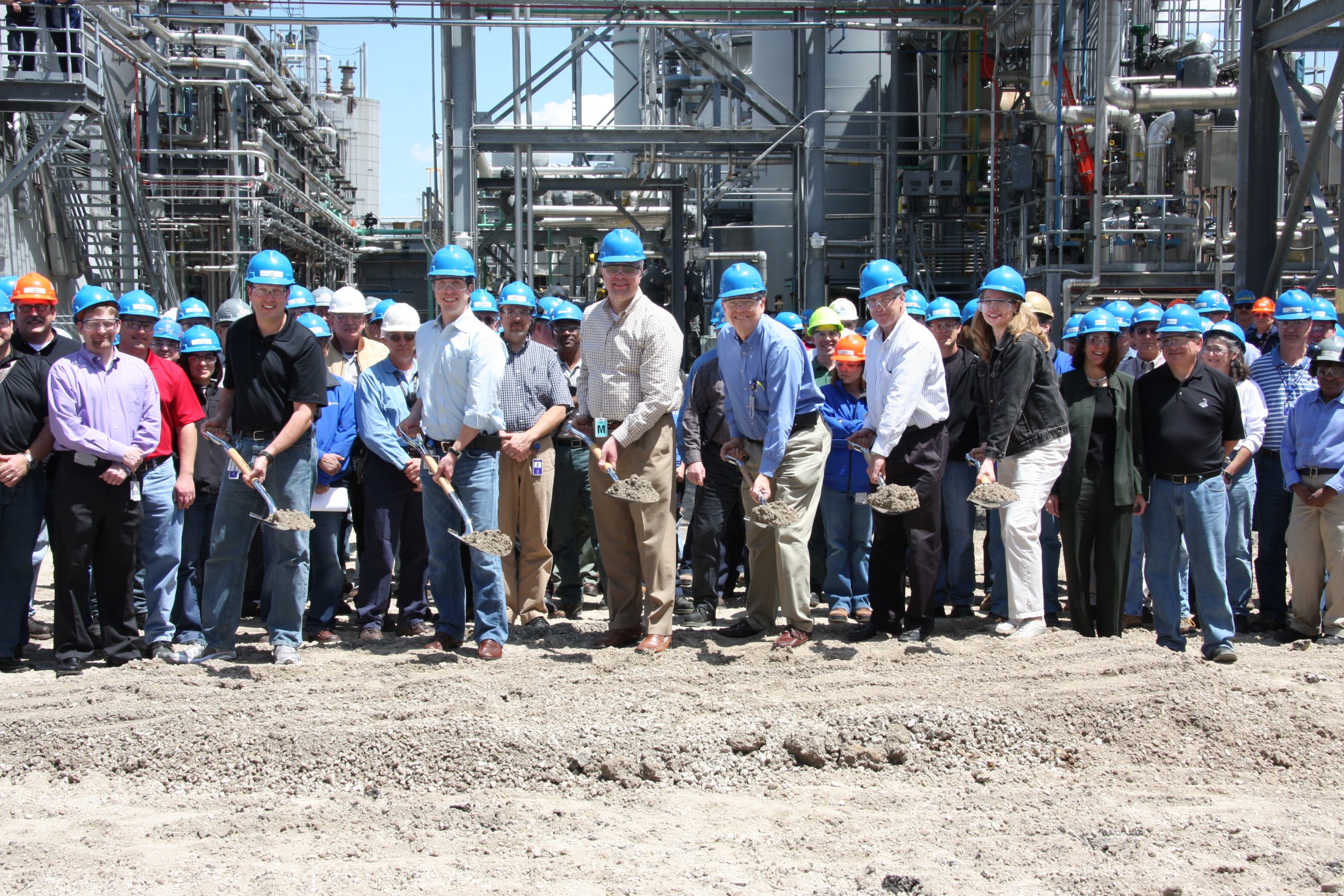 A group of people standing in front of a large industrial plant. They are all wearing blue hard hats and are holding shovels in their hands. The plant appears to be under construction as there are large pipes and equipment visible in the background. The ground is covered in dirt and there is a pile of rubble in the foreground. The people in the group are smiling and appear to be proud of their work.