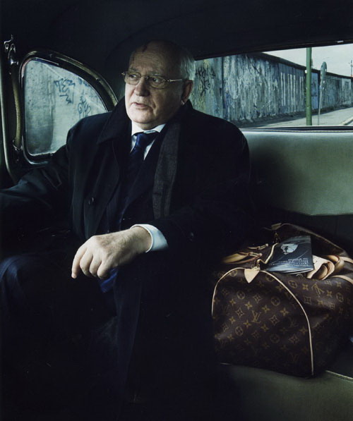 An elderly man sitting in the back seat of a car. He is wearing a black coat a blue tie and glasses. He has a serious expression on his face and is looking directly at the camera. On the right side of the image there is a brown Louis Vuitton handbag with the iconic LV monogram pattern. The car appears to be parked on a street with a concrete wall in the background.