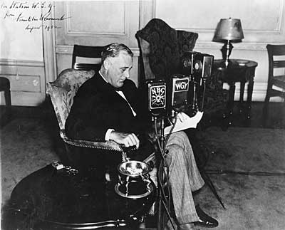 A black and white photograph of a man sitting in a chair in a room with a desk and a lamp in the background. The man is wearing a suit and tie and appears to be in his late twenties or early thirties. He is holding a piece of paper in his hand and is looking down at it with a serious expression on his face. On the desk in front of him there is a microphone with the letters "ABC" and "WGY" on it indicating that he is being interviewed. The photograph appears to have been taken in the early 20th century.