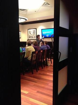 A group of people sitting on wooden chairs in a room with a wooden floor. The room appears to be a conference room or a meeting room as there is a large screen on the wall in the background. The people are gathered around a table with a laptop and other items on it and there are several framed pictures hanging on the walls. The image is taken from a low angle looking up at the people and the screen. The overall mood of the image is casual and relaxed.