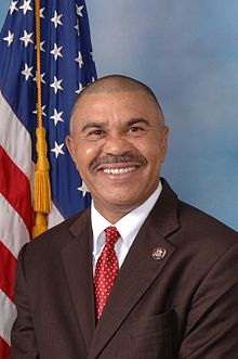 A portrait of a man standing in front of an American flag. The man is wearing a dark suit with a red tie and a white shirt. He has a mustache and is smiling at the camera. The background is a plain blue color. The American flag is visible on the left side of the image.