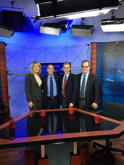 A group of five people standing in front of a news studio set. The set has a blue and red color scheme with a large screen in the background. The people are dressed in formal business attire with three men and two women. They are all smiling and appear to be posing for a photo. In front of them there is a large wooden table with red coffee mugs on it. The studio has a desk and chairs and there are several spotlights hanging from the ceiling.