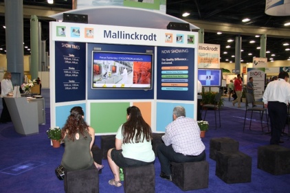 A trade show booth with a large screen on the wall. The booth is called Mallinckrodt and it is located in an exhibition hall with other booths and people in the background. There are four people sitting on black stools in front of the booth looking at the screen. On the screen there is an image of a building and some text that reads "Now Showing". The booth has a blue and white color scheme and there are several potted plants on the floor.