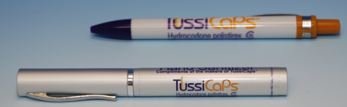 Two pens one in white and the other in orange. The white pen has the brand name "TussiCaps" written on it in black letters. The orange pen has a black cap and a silver body. Both pens have a silver clip attached to the body. The background is a light blue color. The pens appear to be new and unused.