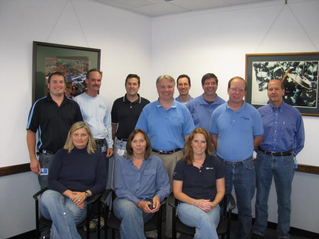 A group of nine people posing for a photo in a room with white walls and a projector screen. There are nine people in total nine men and one woman all wearing blue polo shirts. They are all smiling and looking at the camera. On the left side of the image there is a man wearing a black polo shirt and a woman wearing a blue shirt. In the background there are two framed pictures hanging on the wall. The room appears to be an office or a conference room.