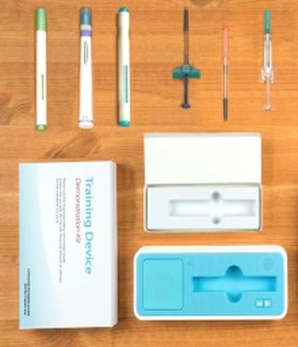A set of medical equipment including a training device a syringe a pen and a box. The training device is white with a blue cover and has the words "Training Device" written on it. There are six pens in total three of which are white with green caps and the other two are blue caps. The syringe is a small cylindrical device with a needle at the end. The box is also white and has a blue lid. All the items are arranged on a wooden surface.