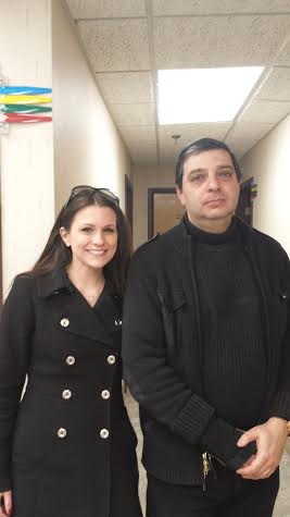 A man and a woman standing in a hallway. The woman is on the left side of the image and the man on the right side. They are both dressed in formal attire with the woman wearing a black coat with gold buttons and a pair of sunglasses on her head. The man is wearing a dark black jacket and a black turtle neck sweater. He has short dark hair and is looking directly at the camera with a serious expression. The hallway has white walls and a tiled floor. There is a colorful banner hanging on the wall in the background.