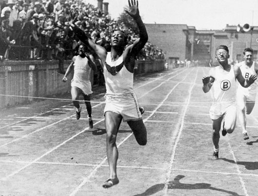 A black and white photograph of a male athlete running on a track. He is in the middle of a race with his arms stretched upwards and his body slightly bent forward. He appears to be in the lead with a determined expression on his face. Behind him there are two other male athletes one on the right side of the image and the other on the left side who are also running towards him. The athlete in the foreground is wearing a white tank top and shorts and his arms are raised in triumph. The background shows a large crowd of spectators in the stands and a building can be seen in the distance. The image appears to have been taken at a sporting event possibly a track and field event.