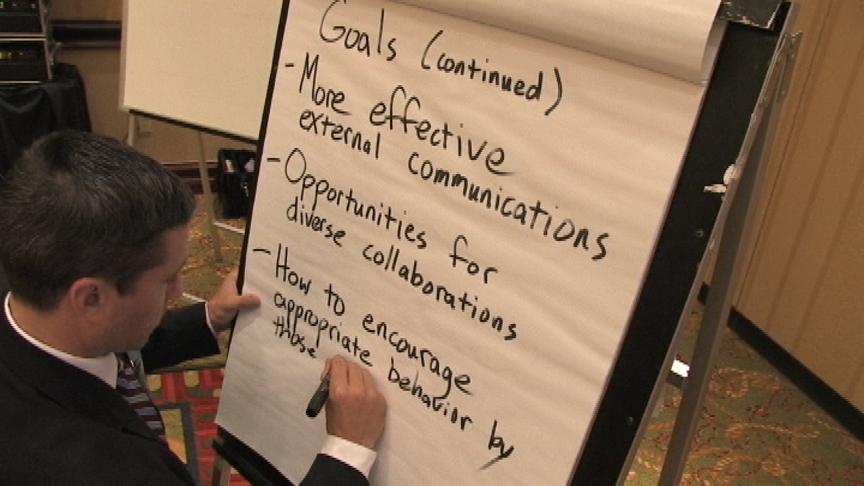 A man in a suit writing on a whiteboard. He is standing in front of a projector screen and appears to be giving a presentation. The whiteboard has a list of goals written on it in black marker. The goals are:<br /><br />- Goals (Continued)<br />- More effective external communications<br />- Opportunities for diverse collaborations<br />- How to encourage appropriate behavior<br /><br />The man is holding a marker in his right hand and is writing on the whiteboard with it. He seems to be focused on the task at hand. The background of the image is a conference room with a carpeted floor and a projector.