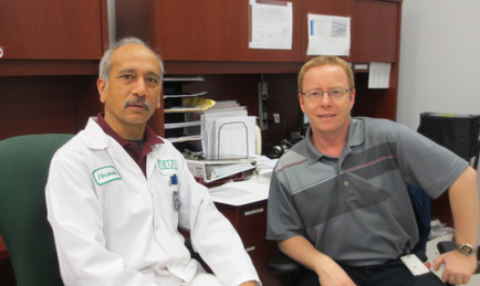 Two men sitting in an office. The man on the left is wearing a white lab coat with a green logo on it and has a mustache. He is sitting on a green office chair and is looking directly at the camera with a serious expression on his face. On the right is a man wearing a gray polo shirt and glasses. He has short blonde hair and is sitting in front of a desk with papers and other office supplies scattered around. The background shows a wooden cabinet and a computer monitor.
