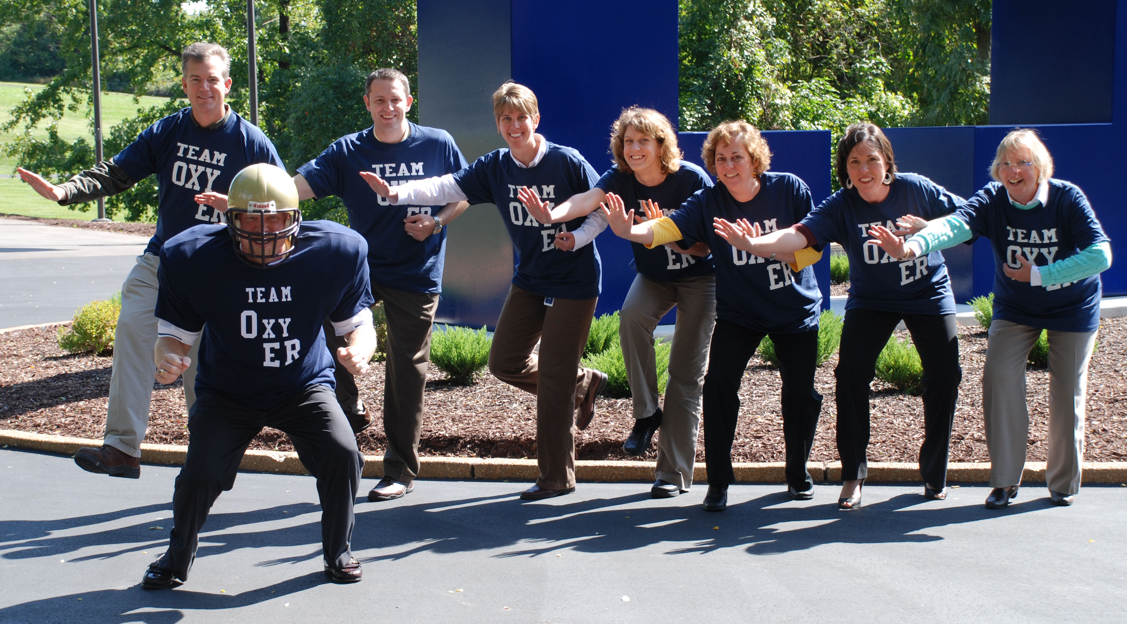 A group of eight people standing in front of a blue structure with trees and bushes in the background. They are all wearing blue t-shirts with the words "Team OXY ER" printed on them. Seven of the people stand in a line in the background appearing to pretend like they are throwing a football. A man at the front of the photograph is wearing full football gear and squatting. The group appears to be posing for a photo.