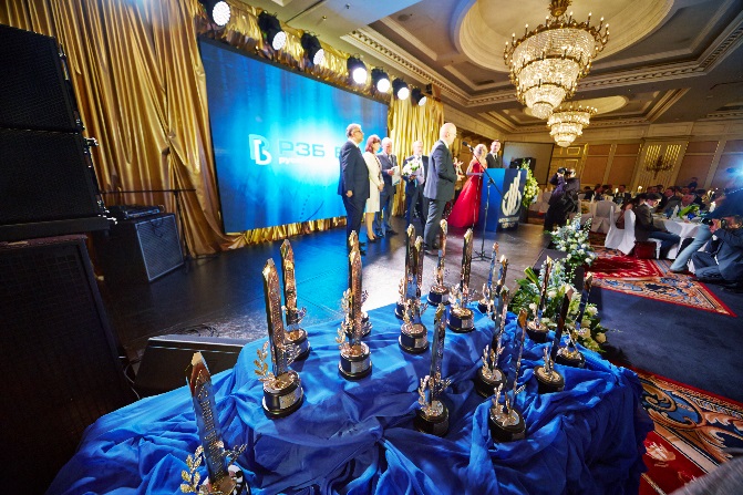 A large room with a high ceiling and a chandelier hanging from it. The room is decorated with gold curtains and a large screen in the background. On the stage there are several people standing and talking to each other. In the center of the room there is a table covered with a blue tablecloth and several golden trophies. The trophies are arranged in a row and appear to be of different sizes and shapes. There are also a few people sitting at tables on either side of the stage. The overall atmosphere of the image is formal and celebratory.