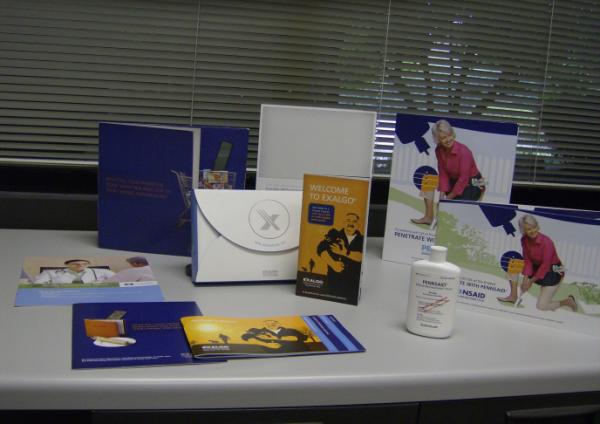 A desk with a variety of items on it. There are six items on the desk including a blue folder a white envelope a brochure a book a bottle of hand sanitizer and a few pamphlets. The items are arranged neatly on the table and there is a window with blinds in the background. The desk appears to be in an office or a workspace as there are no other items visible in the image.