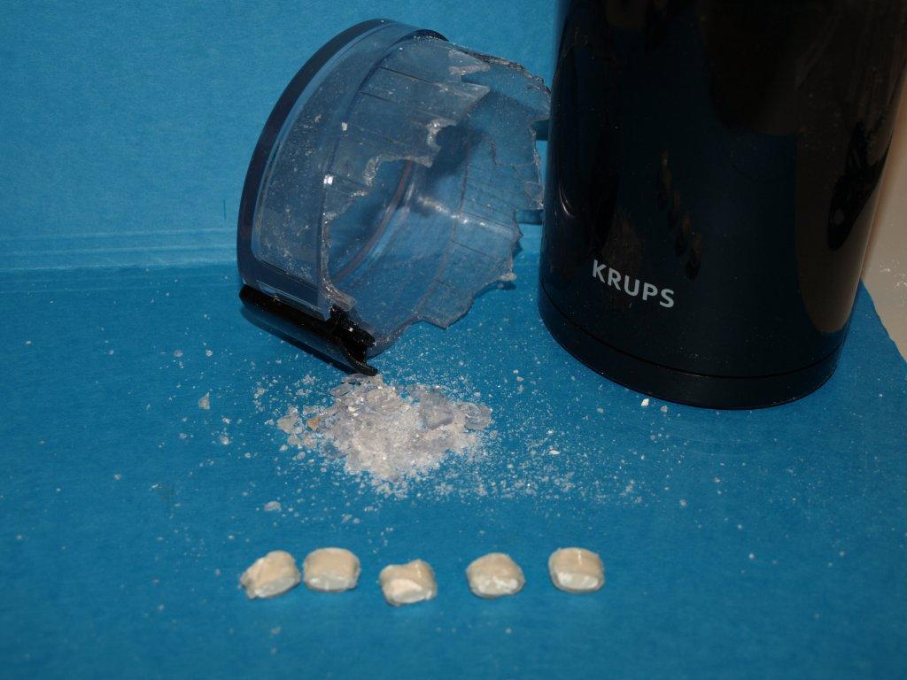 A black Krups coffee grinder on a blue surface. The grinder has a black body with the word "Krups" written on it in white letters. Next to the grinder there is a small pile of white powder which appears to be sugar or salt. There are also a few small white pills scattered around the pile. The background is blurred but it seems to be a kitchen countertop.