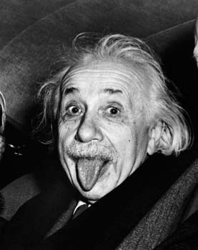 A black and white photograph of Albert Einstein the famous physicist and Nobel Prize-winning physicist. He is seen sticking his tongue out in a playful manner with his eyes wide open and his mouth slightly open. His hair is styled in a messy curly manner and he is wearing a suit and tie. The background is blurred but it appears to be an indoor setting with other people visible in the background.