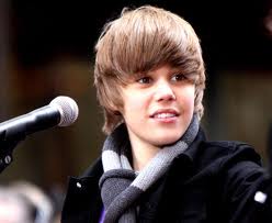 A close-up portrait of a young man who appears to be in his late teens or early twenties. He has blonde hair styled in loose waves and is wearing a black jacket and a gray scarf around his neck. He is standing in front of a microphone and is looking directly at the camera with a slight smile on his face. The background is blurred but it seems like he is on a stage or a public event.