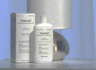 A white bottle of Pennsaid skincare products on a grey surface. The bottle is cylindrical in shape with a white cap and has a label on it. Next to the bottle there is a white box with the brand name "Pennsaid" written in black text. The box is rectangular in shape and appears to be made of cardboard. The background is a light blue color. On the right side of the image there are two silver lamps with white shades. The lamps are placed next to each other creating a modern and minimalist look.