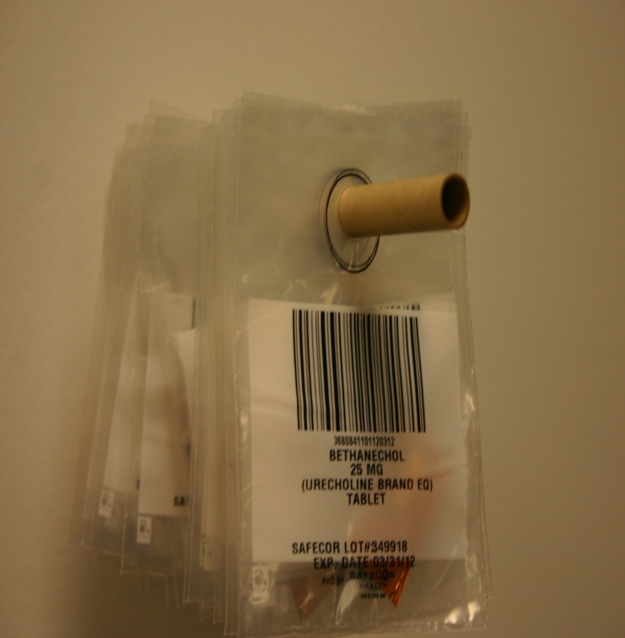 A transparent plastic bag with a label attached to it. The label has a barcode and text that reads "BETHANCHOL (URCHOLINE BRAND EO) TABLET". The label also has a small wooden handle attached to the top right corner of the bag. The bag appears to be sealed and is hanging on a white wall.