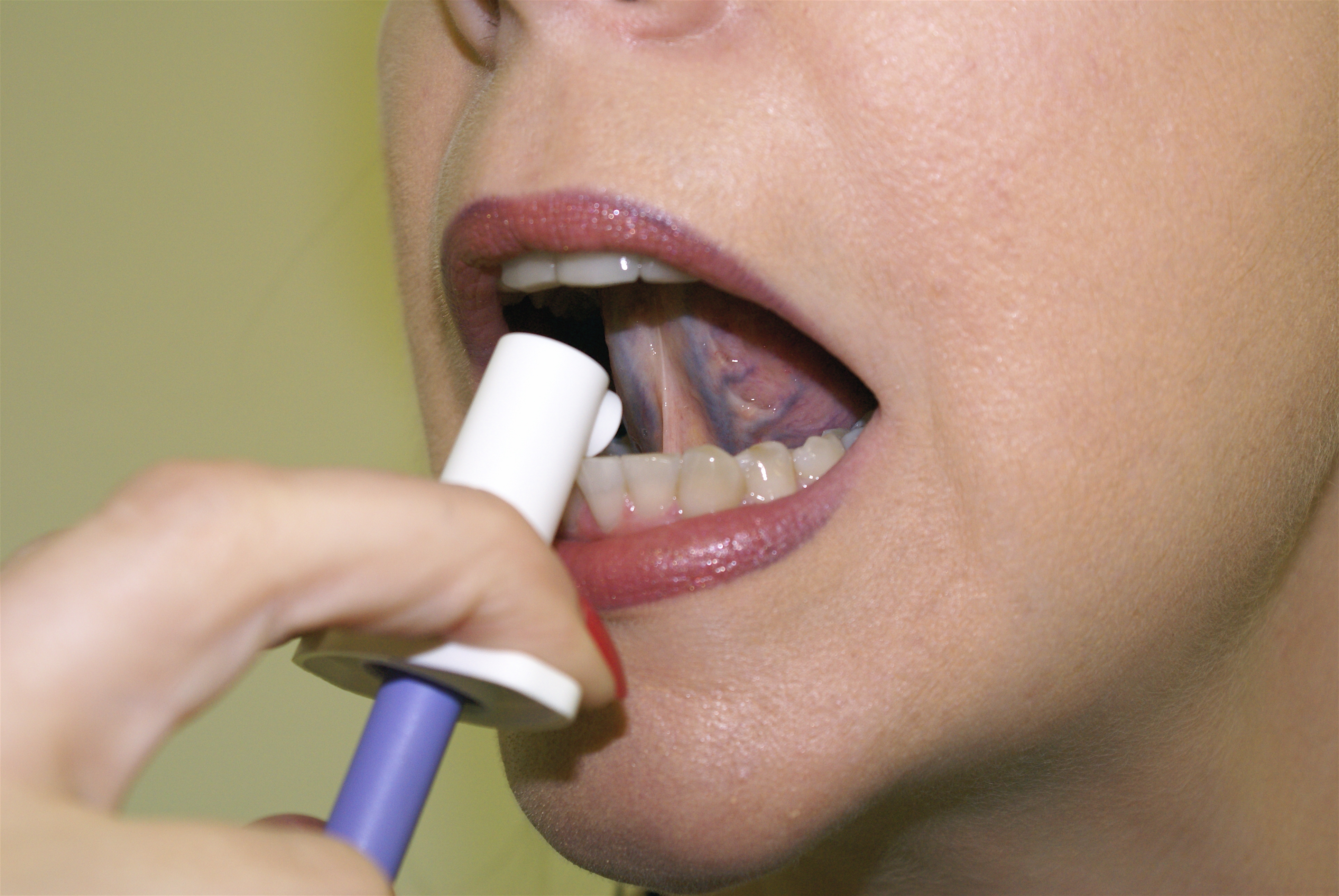 A close-up of a person's mouth with their mouth open and their tongue sticking out. The person is holding a blue and white toothbrush in their right hand and is using it to clean their teeth. The toothbrush has a white cap and a blue handle. The background is a plain yellow color. The image appears to be taken in a dental office or clinic setting.