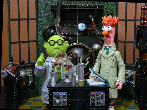 Two characters from "The Muppets" standing in front of a large machine. They are Dr. Bunsen Honeydew and Beaker. Dr. Bunsen Honeydew is a green man with glasses and a white lab coat pointing his index finger. He is standing next to a black table with various scientific equipment on it. On the right is Beaker a pink man with orange hair and a green suit. He has a surprised expression on his face. In the background there is a large black machine with pipes and valves. The room appears to be a laboratory or laboratory setting with a green and yellow checkered floor.